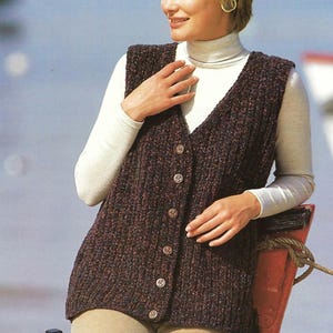 May include: A brown, button-down vest with a V-neck, worn over a white turtleneck. The vest has a textured knit pattern and is paired with tan corduroy pants. The background shows a body of water.