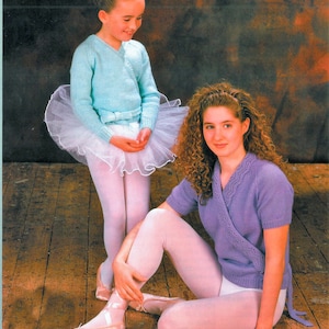 May include: Two girls wearing ballet-inspired knit sweaters. The younger girl is wearing a light blue sweater with a white tutu and white tights. The older girl is wearing a purple sweater with white tights. Both girls are wearing ballet shoes.