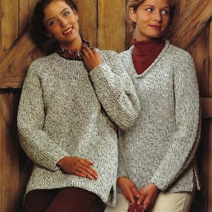 May include: Two women in speckled grey knit sweaters. One sweater has a crew neck, the other a V-neck. They stand before a wooden door. One wears a patterned scarf, the other a red turtleneck. The sweaters appear to be made of a thick yarn.