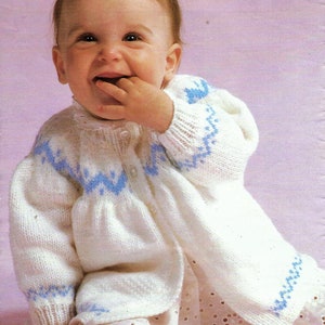 May include: A baby wearing a white knitted cardigan with blue and white patterned trim. The cardigan has a button closure and is worn over a white lace dress.