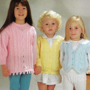 May include: Three children wearing knitted sweaters in pink, yellow, and blue. The sweaters feature a cable knit pattern. The girl on the left is wearing a pink sweater with a scalloped hem. The girl in the middle is wearing a yellow sweater with a V-neck. The girl on the right is wearing a blue vest with buttons.