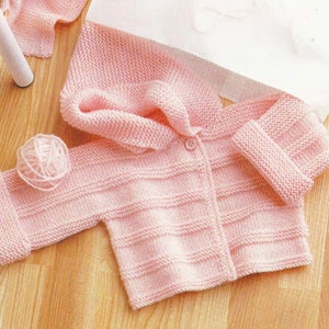May include: A pink knitted baby cardigan with a hood and button fastening. The cardigan has a ribbed texture and is made with soft yarn.