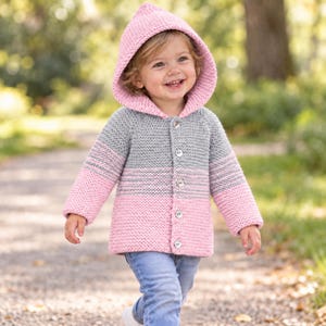 May include: A hooded knit cardigan featuring a colour-blocked design with a soft pink hood and hem, a grey midsection, and light pink sleeves. The garment is secured with five round buttons down the front and is crafted with a textured garter stitch.