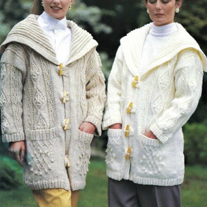 May include: Two women wearing cream-colored knitted cardigans with large collars and wooden toggle closures. The cardigans have a cable knit pattern and pockets.