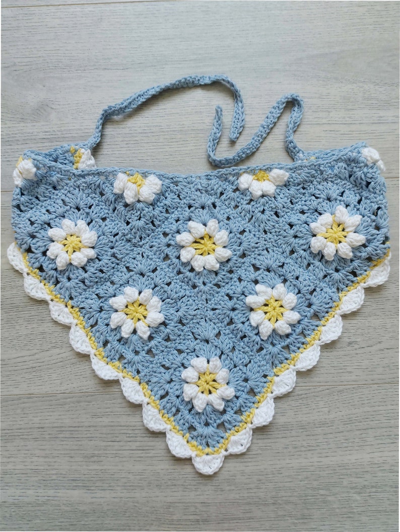 Puede incluir: Una bufanda triangular de crochet azul claro con un patr&oacute;n de margarita blanca y amarilla. La bufanda tiene un borde festoneado y se ata al cuello.