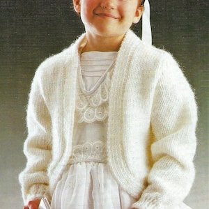 May include: A white knitted cardigan with long sleeves and a shawl collar. The cardigan is cropped and has a slightly fitted silhouette. The model is wearing a white dress with lace trim underneath the cardigan.