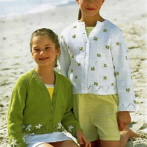 May include: Two girls wearing knitted cardigans. The girl on the left is wearing a green cardigan with white embroidered flowers. The girl on the right is wearing a white cardigan with green embroidered flowers. Both girls are sitting on a sandy beach.