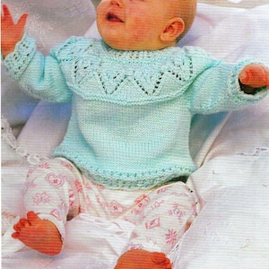 Puede incluir: Un bebé con un suéter de punto azul claro con un escote de encaje. El suéter tiene un diseño simple y es perfecto para el primer invierno de un bebé.