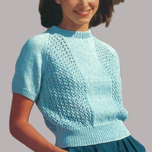 May include: A light blue knit top with short sleeves and a textured openwork design on the front. The top has a ribbed hem and neckline. The person is wearing a teal skirt. The overall style is casual and comfortable.