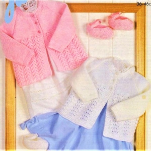Puede incluir: Dos cárdigans de punto para bebé, uno rosa y otro blanco, con cierre de botones y un delicado patrón de encaje. Los cárdigans se muestran sobre un fondo blanco con un marco de madera.