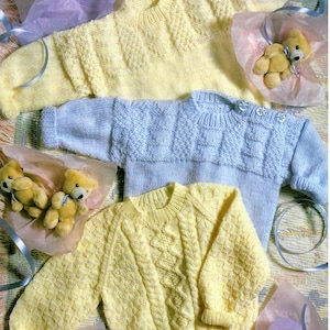 Könnte beinhalten: Drei gestrickte Baby-Pullover in Pastellgelb und Blau. Die gelben Pullover haben Rauten- und Zopfmuster. Der blaue Pullover hat ein strukturiertes Design. Jeder Pullover hat lange Ärmel und ist mit kleinen Knöpfen verziert. Teddybären sind im Bild enthalten.