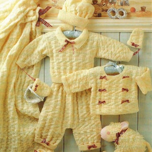 May include: A knitted baby outfit set in a pale yellow colour with a matching hat, booties, and a stuffed lamb. The set features a cardigan, trousers, and a blanket with red and white ribbon accents.