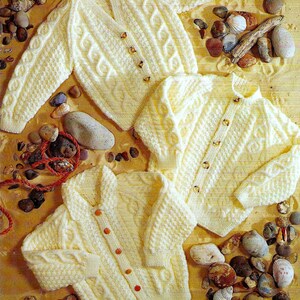 May include: Three white knitted cardigans with button closures, featuring a cable knit pattern. The cardigans are arranged on a background of tan fabric with small stones and seashells.