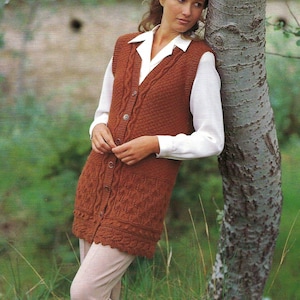 Puede incluir: Un chaleco largo de punto color óxido con botones, usado sobre una camisa blanca con cuello. El chaleco presenta un patrón de punto texturizado y se combina con pantalones beige. La persona está apoyada en un árbol en un entorno natural.