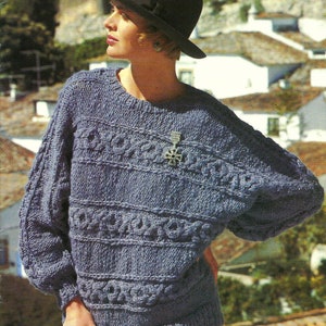 Op de afbeelding: Een blauwe gebreide trui met een kabelbreipatroon en een wijde, losse pasvorm. De trui heeft een ronde hals en lange mouwen. Het model draagt een zwarte hoed.