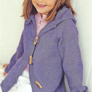 May include: A young person wearing a purple knitted hooded cardigan with wooden toggle closures. The cardigan is paired with a pink shirt and white pants. The person is smiling.