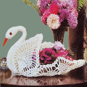 May include: A white crocheted swan-shaped basket filled with pink and purple flowers. The swan has a red beak and eye details. The basket is open, displaying the flowers. The background includes a vase of flowers.