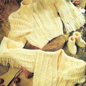 May include: Two knitted baby cardigans with fringe trim and matching booties. The cardigans are cream colored and have a chevron pattern. The booties are also cream colored and have a simple knit pattern.