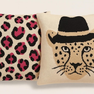 May include: Two decorative throw pillows. One features a cream background with pink and black leopard spots. The other pillow has a cream background with a cheetah face wearing a black hat. The pillows are square.
