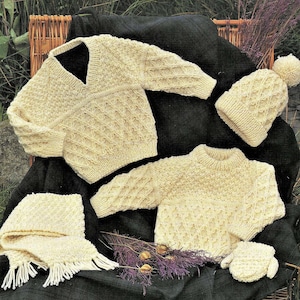 May include: A set of knitted baby clothes in a cream colour, including a sweater, hat, mittens, and scarf. The items are laid out on a black and white checked fabric.