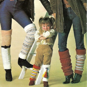 May include: Three people are wearing leg warmers. The person in the middle is a young girl with pigtails. She is wearing a brown vest and brown pants. The leg warmers are white with colorful tassels, red, and striped.