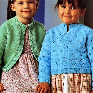 May include: Two young girls wearing knitted cardigans. The girl on the left is wearing a green cardigan with buttons. The girl on the right is wearing a blue cardigan with a diamond pattern.