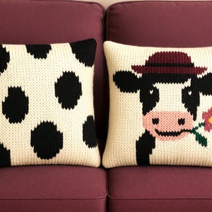 May include: Two decorative throw pillows with a cow theme. One pillow features black spots on a cream background. The other pillow has a cow wearing a hat and holding a flower. Both pillows are on a purple couch.