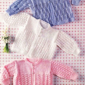May include: Three knitted baby cardigans in different colours: light blue, white and pink. Each cardigan has a button fastening and a delicate lace pattern.