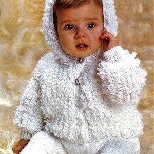 May include: A white knitted baby cardigan with a hood, featuring a textured, bobbly stitch. The cardigan has a button closure and is worn by a baby with brown hair.