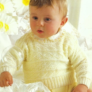 Könnte beinhalten: Ein Baby trägt einen gelben Strickpullover mit weißem Kragen. Der Pullover hat ein strukturiertes Muster und das Baby sitzt auf einer weißen Spitze.