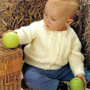 May include: A baby wearing a white knitted cardigan with buttons and blue jeans sits on a straw bale holding a green apple.