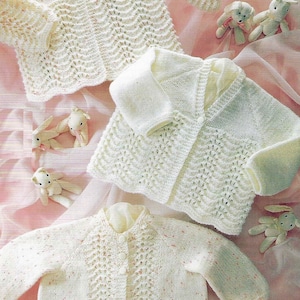 Op de afbeelding: Drie witte gebreide baby-vestjes met knopen. De vestjes liggen op een roze achtergrond met kleine witte knuffels.