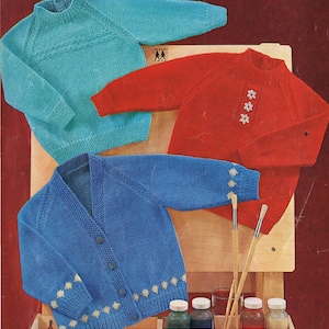 May include: Three knitted sweaters in different colors: a teal blue sweater, a red sweater with white flower embroidery, and a blue cardigan with white snowflake embroidery. The sweaters are displayed on an easel with paintbrushes and paint jars.
