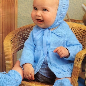 May include: A baby wearing a blue knitted cardigan, matching booties, and a bonnet with a lace pattern. The baby is sitting in a wicker chair.