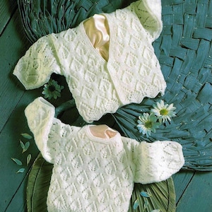May include: Two white crocheted baby sweaters with a delicate lace pattern. One sweater has a V-neck and the other has a round neck.