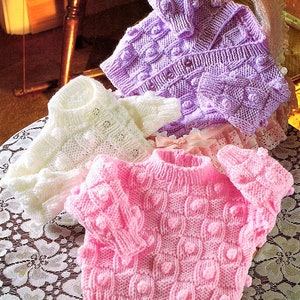 Könnte beinhalten: Drei gestrickte Baby-Pullover in Rosa, Weiß und Lila mit einer strukturierten Noppenmasche. Die Pullover liegen auf einer weißen Spitzentischdecke.