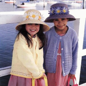 May include: Two girls wearing knitted cardigans and straw hats decorated with flowers. The girl on the left is wearing a yellow cardigan and a hat with yellow daisies. The girl on the right is wearing a purple cardigan and a hat with purple daisies.