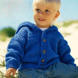 Può includere: Un cardigan blu lavorato a maglia con cappuccio e bottoni in legno. Il cardigan è indossato da un bambino piccolo. L'indumento ha orlo e polsini a coste. Il bambino indossa pantaloni blu e una camicia bianca sotto il cardigan.
