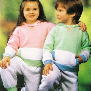May include: Two children wearing knitted sweaters and pants. The girl is wearing a pink and white sweater with green trim and white pants. The boy is wearing a green and white sweater with blue trim and white pants.