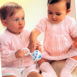 Puede incluir: Dos bebés con atuendos de punto rosa. Un bebé lleva un cárdigan y el otro un vestido. Ambos bebés llevan calcetines rosas. El bebé con el cárdigan sostiene un sonajero azul y blanco.