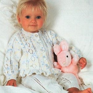 May include: A baby wearing a white crocheted cardigan with blue and yellow accents and matching trousers. The cardigan has a ruffled hem and the baby is holding a pink plush bunny.