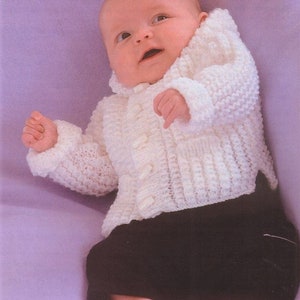 May include: A baby wearing a white knitted cardigan with buttons and black trousers. The baby is lying on a purple blanket.