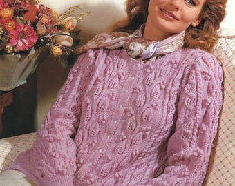 May include: A light purple knitted sweater with a detailed leaf and bobble pattern. The sweater has long sleeves and a ribbed hem and cuffs. A patterned scarf is draped around the neck.