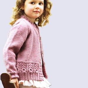 Puede incluir: Una niña pequeña con un cárdigan de punto rosa con un delicado ribete de encaje. El cárdigan tiene cierre de botones y mangas largas. La niña tiene el pelo rubio rizado y lleva una camisa de rayas blancas y rosas debajo del cárdigan.