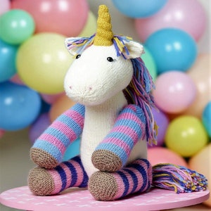 May include: A white knitted unicorn toy with pink and blue striped legs and a yellow, blue, and purple mane and tail. The unicorn is sitting on a pink surface.