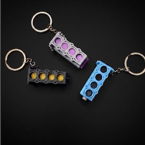 May include: Three engine block keychains with metal rings. One is black with yellow circular inserts, one is silver with purple circular inserts, and one is light blue with dark blue circular inserts. Each keychain features four cylinder openings.