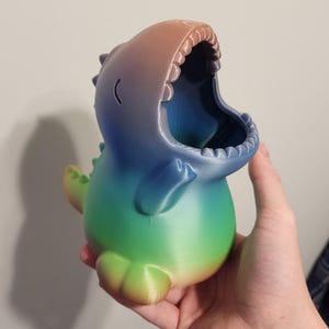 May include: A 3D-printed dinosaur-shaped container with a wide open mouth. The container is a gradient of colors, transitioning from green to blue to orange. The dinosaur has small arms and a curved line for an eye.