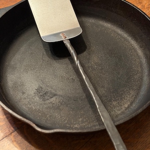 Può includere: Una spatola in acciaio inossidabile con manico attorcigliato poggia su una padella in ghisa nera. La spatola ha una testa rettangolare e un manico scuro e strutturato. La padella è rotonda e ha una finitura scura e opaca.