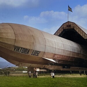 May include: A large, brown airship, the "Viktoria Luise", is being pulled out of a hangar. The airship is partially visible, with the text "Viktoria Luise" on the side. There are people standing around the airship and hangar.