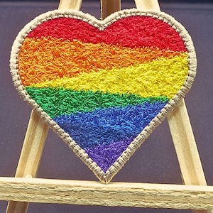 May include: A rainbow heart-shaped patch with a fuzzy texture. The patch is made of red, orange, yellow, green, blue, and purple fabric.